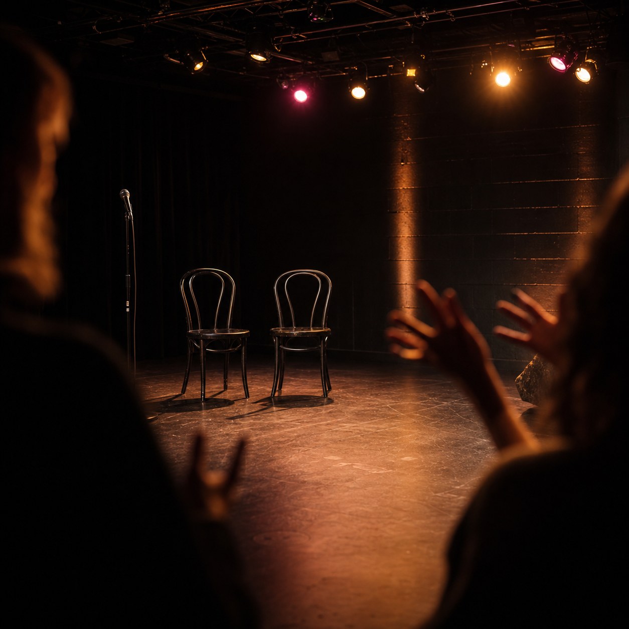 Warm black-box theater stage with two chairs, a microphone, and a blurred audience.