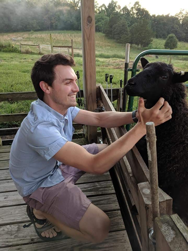 Ben smiling while crouched beside a black sheep on a farm.